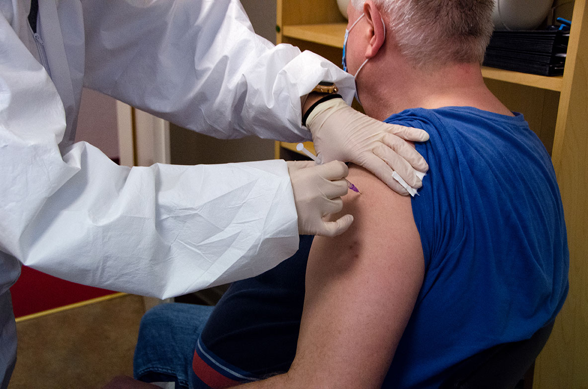 Das Foto zeigt eine Medizinische Fachangestellte, die einem vor ihr sitzenden Patienten eine Spritze in den Oberarm gibt. Der Patient trägt eine Atemschutzmaske.
