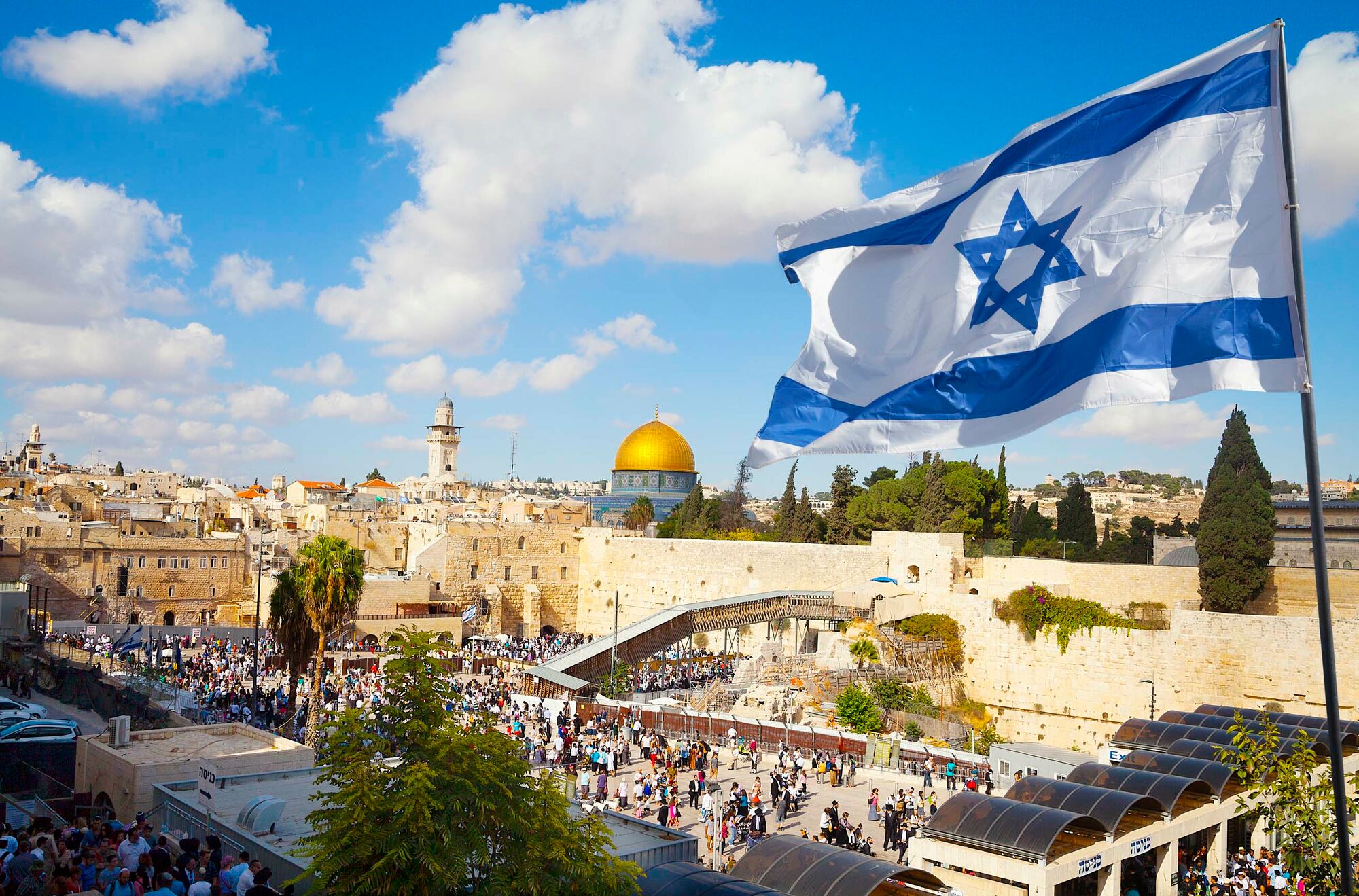 Das Foto zeigt einen Teil der Stadt Jerusalem mit der Klagemauer im Vordergrund. Rechts im Bildvordergrund weht eine israelische Flagge.