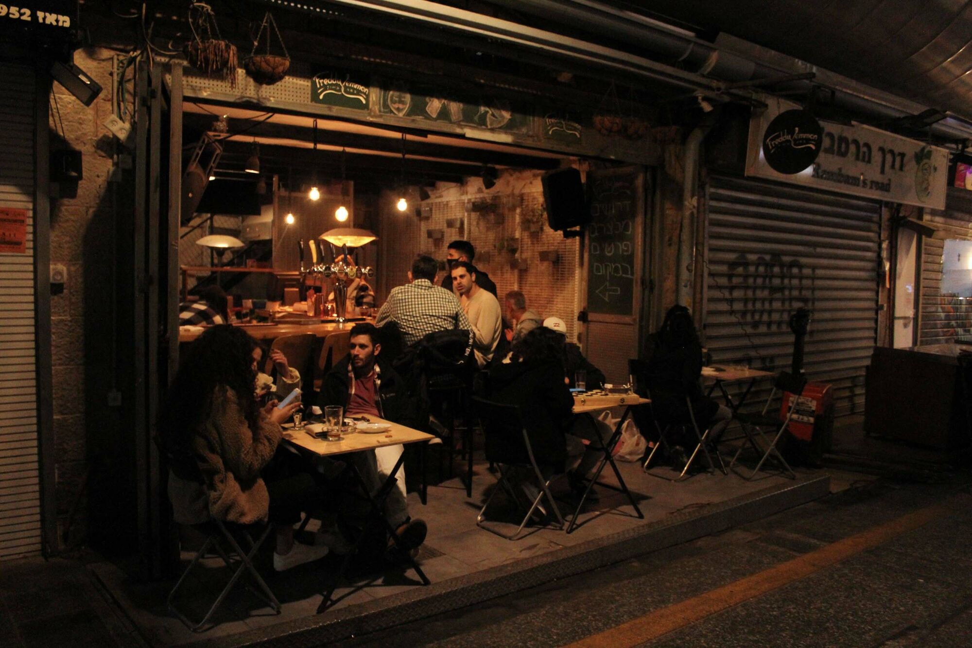 Das Foto zeigt eine kleine Bar in Israel am Abend. Vor der Bar stehen drei Tische auf der Straße. Am Tresen und an den Tischen sitzen Gäste.