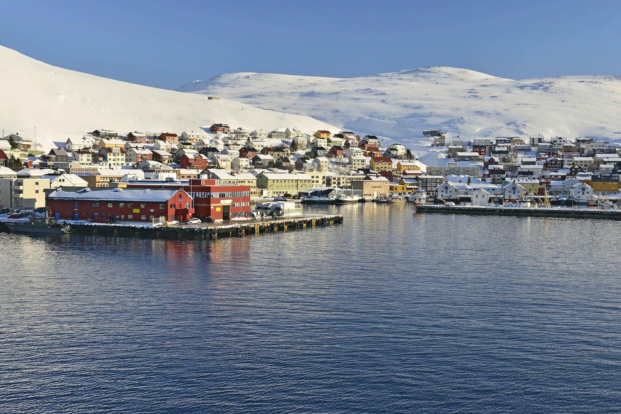 Das Foto zeigt die kleine Stadt Honningsvåg im Norden Norwegens vom Wasser aus gesehen. Hinter der Stadt sind schneebedeckte Berge zu sehen.