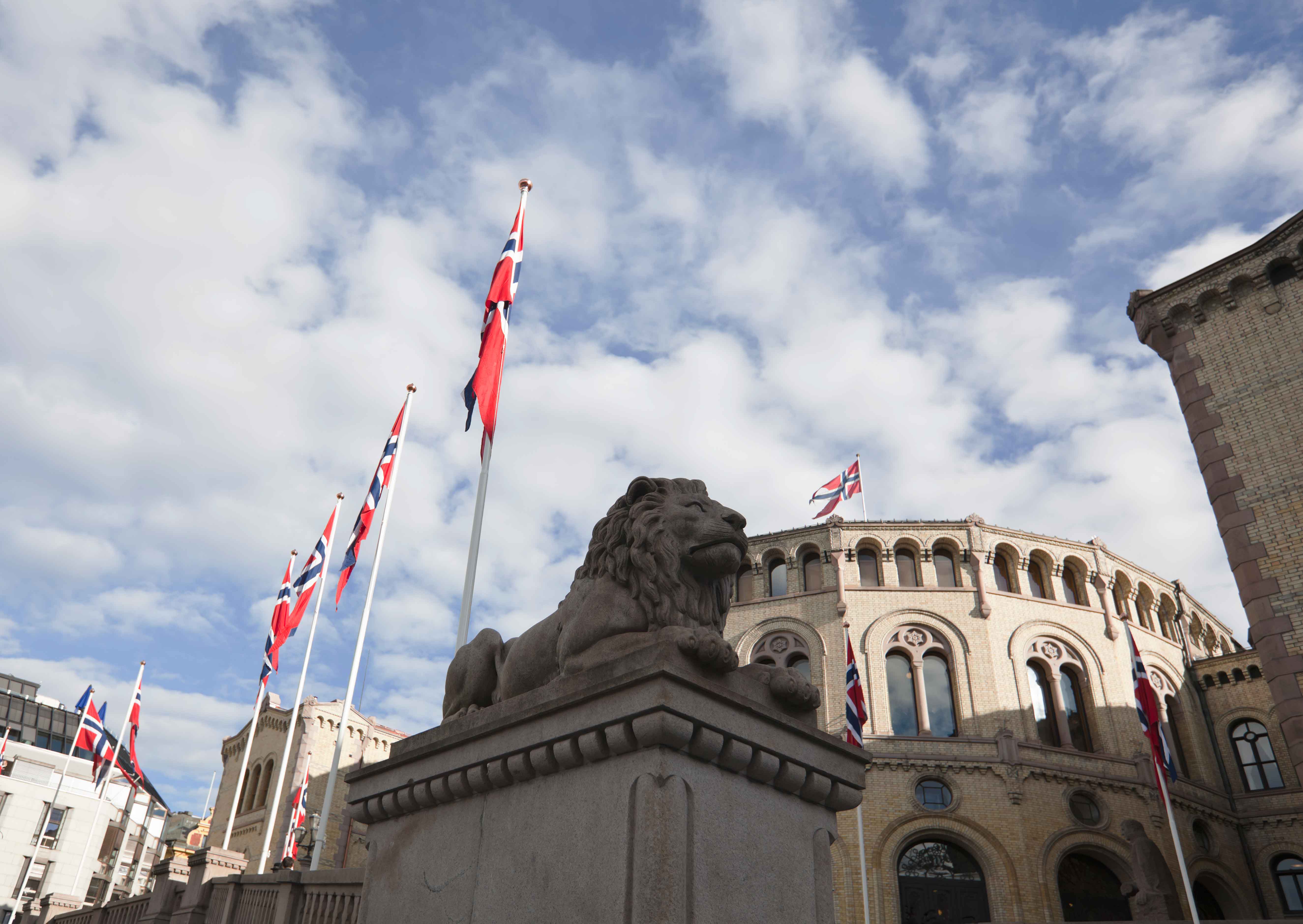 Das Foto zeigt das norwegische Parlament in Oslo. Vor dem Gebäude stehen Fahnenmasten mit norwegischen Flaggen.