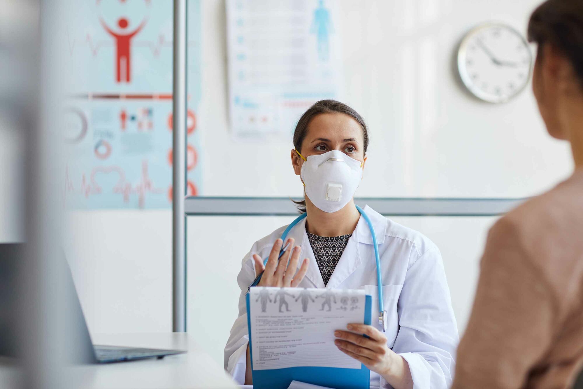 Das Foto zeigt eine Ärztin in weißem Kittel mit Stethoskop um den Hals und einem Klemmbrett in der Hand im Gespräch mit einer ihr gegenüberstehenden Person.