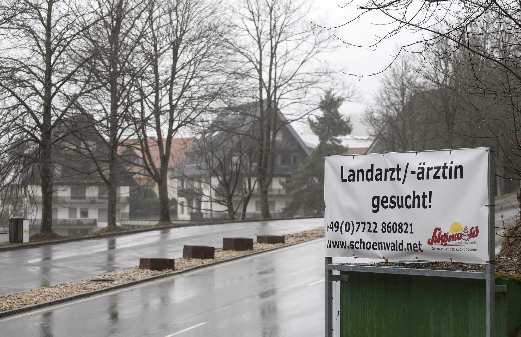 Das Foto zeigt ein Schild aus Planenstoff an einer Straße. Auf dem Schild steht „Landarzt/-ärztin gesucht!“, darunter die Telefonnummer +49 (0)7722 860821, die Internetadresse www.schoenwald.net sowie das Logo des Dorfes Schönwald im Schwarzwald. Im Hinte