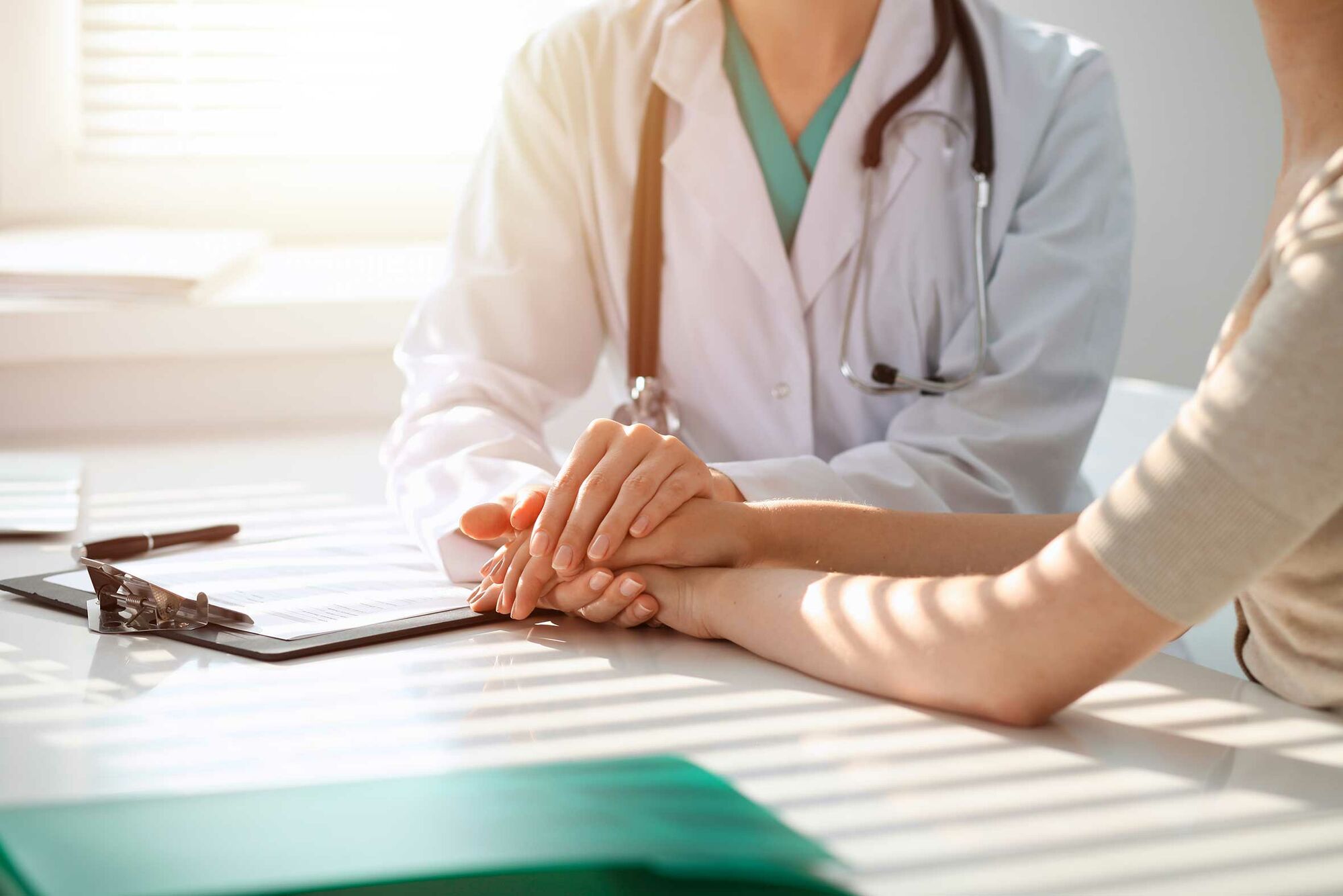 Das Bild zeigt eine Medizinerin in weißem Kittel mit Stethoskop um den Hals, die mit einer Patientin an einem Tisch sitzt und deren Hände umfasst. Vor der Medizinerin liegt ein Klemmbrett auf dem Tisch.