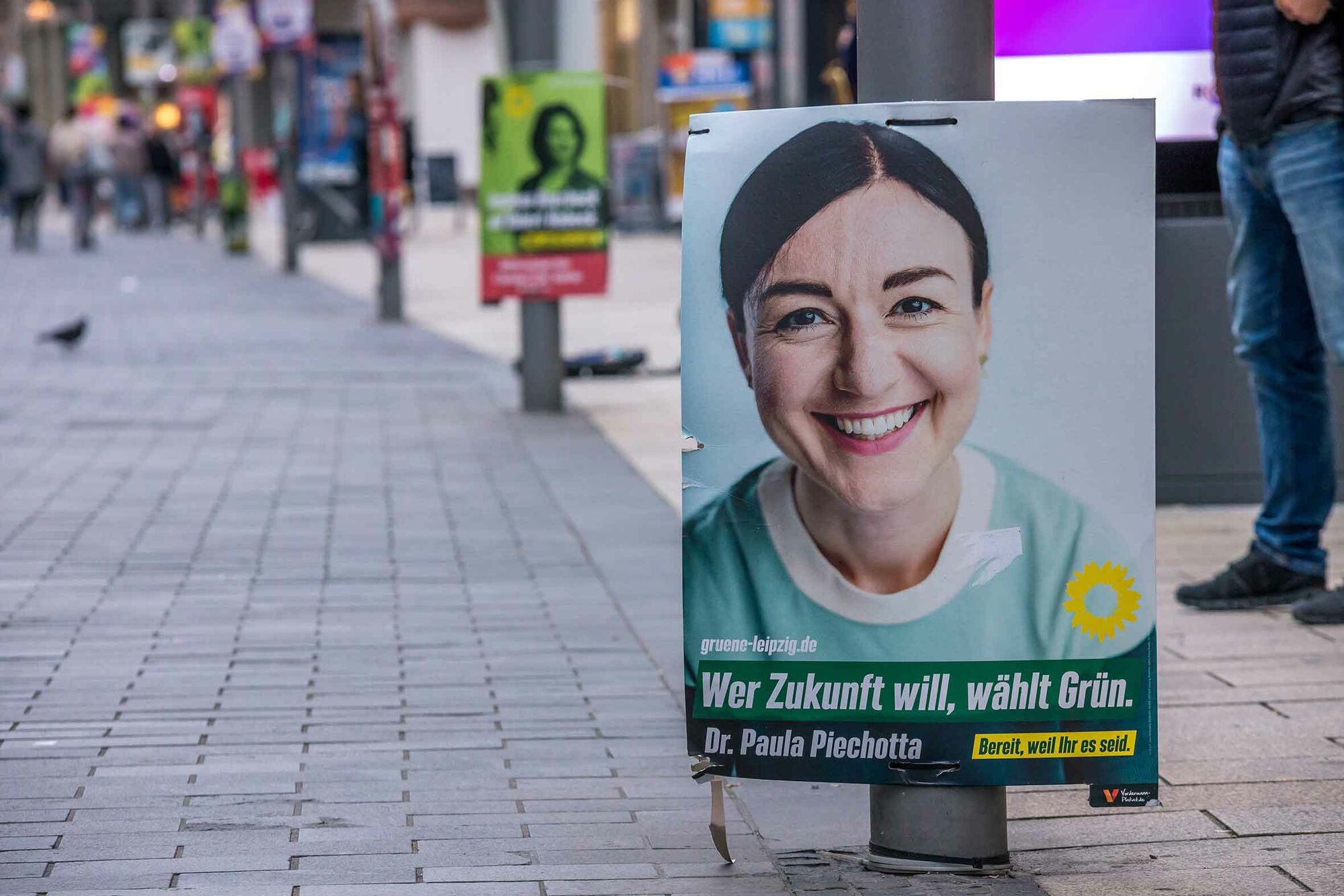 Das Foto zeigt eine Fußgängerzone, in der ein Wahlkampfplakat der Grünen mit dem Gesicht Paula Piechottas zu sehen ist. Auf dem Plakat steht: Wer Zukunft will, wählt Grün. Dr. Paula Piechotta. Bereit, weil Ihr es seid: grünen-leipzig.