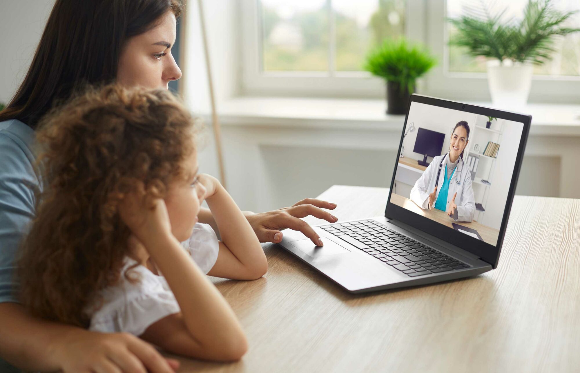 Das Foto zeigt eine Frau mit Kind, die zu Hause vor einem Laptop sitzen und während einer Videosprechstunde den Ausführungen einer Ärztin lauschen.