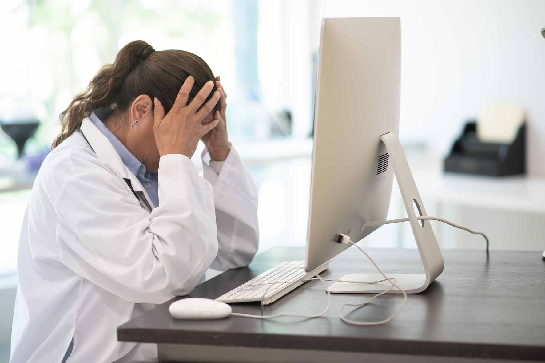 Das Foto ist ein Symbolbild, auf dem eine Ärztin vor ihrem Computer sitzt und die Hände über dem Kopf zusammenschläft.