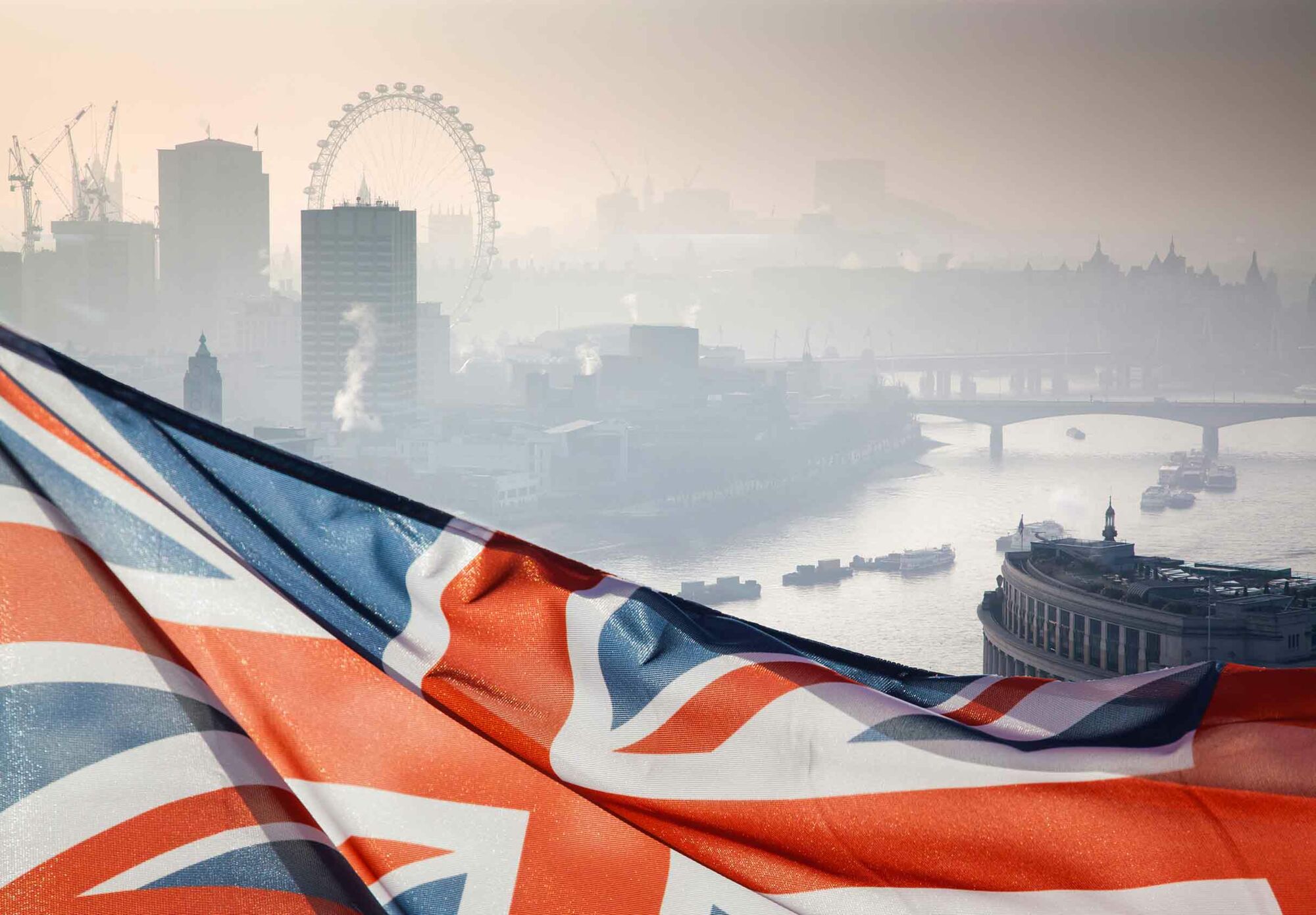 Das Foto zeigt London und die Themse im Nebel. Im Vordergrund weht der britische Union Jack.