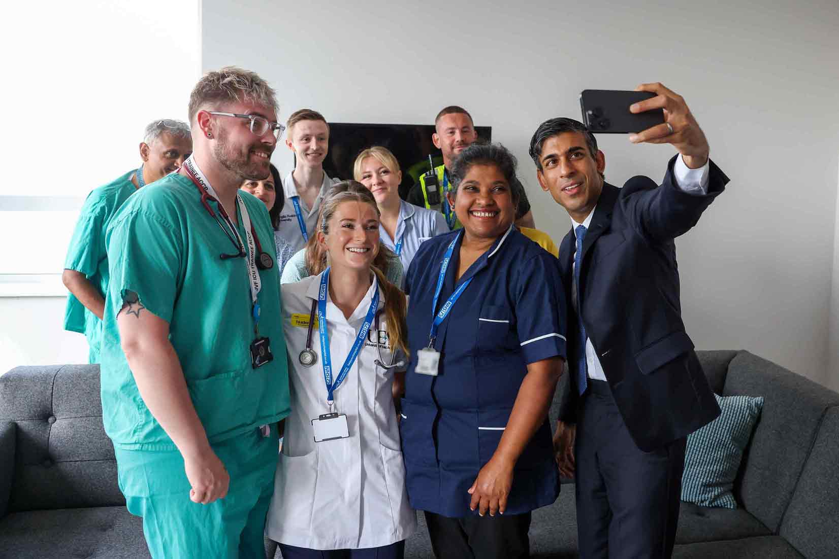Das Foto zeigt den britischen Premierminister Rishi Sunak, während er mit Mitarbeitenden des Addenbrooke's Hospitals in Cambridge ein gemeinsames Selfie macht.