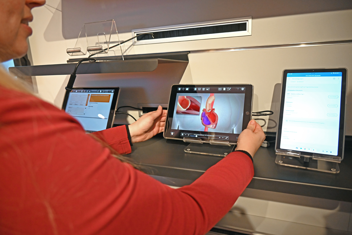 Eine Frau im rotem Pullover erklärt in der Showpraxis der KV Berlin die unterschiedliche Nutzung von digitalen Gesundheitsanwendungen am Beispiel einer Software zur Darstellung von Operationen oder Krankheitsbildern.