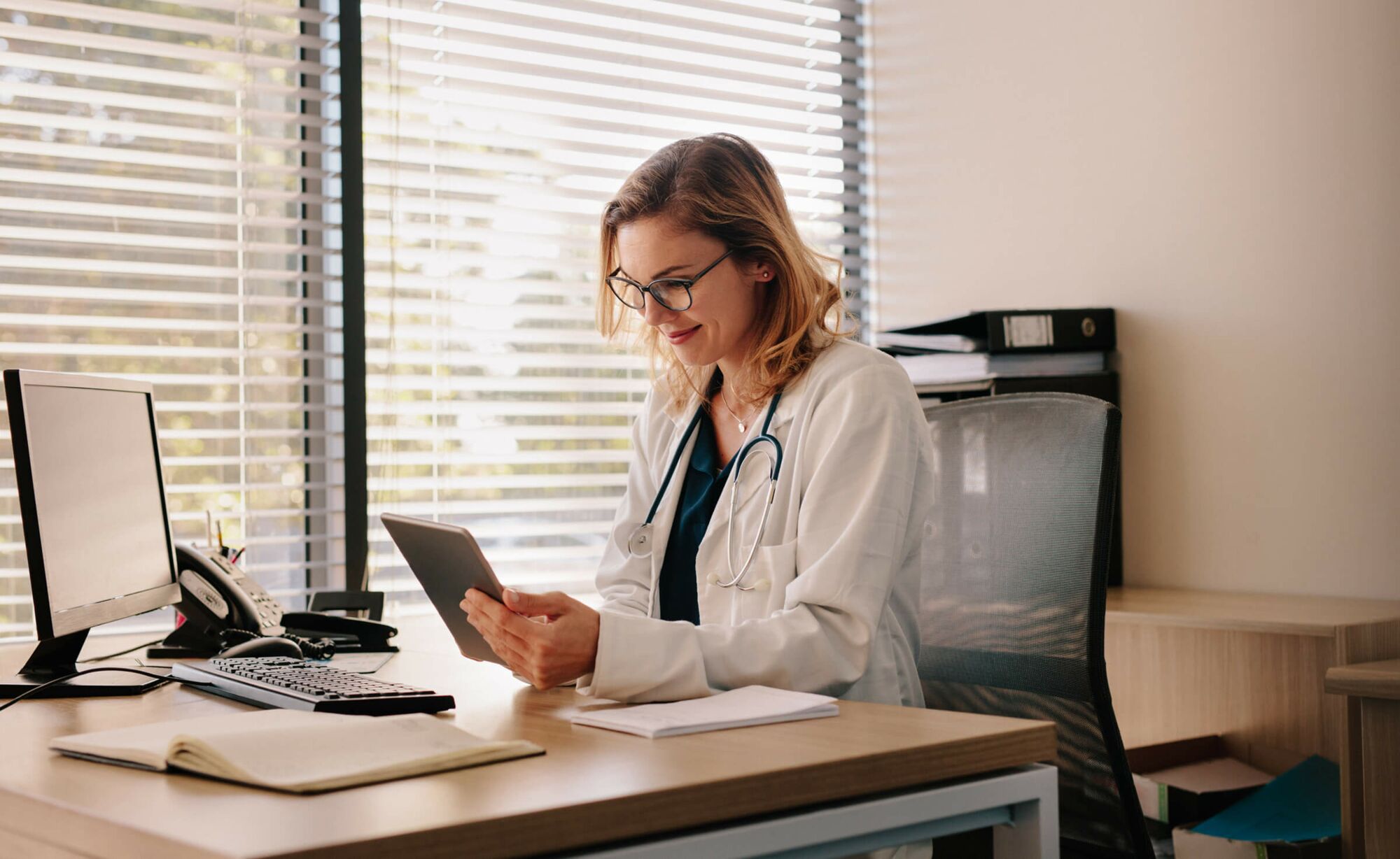 Eine junge blonde Ärztin mit Brille und Stethoskop um den Hals sitzt an einem Schreibtisch und schaut zufrieden auf das Tablet n ihrer Hand.
