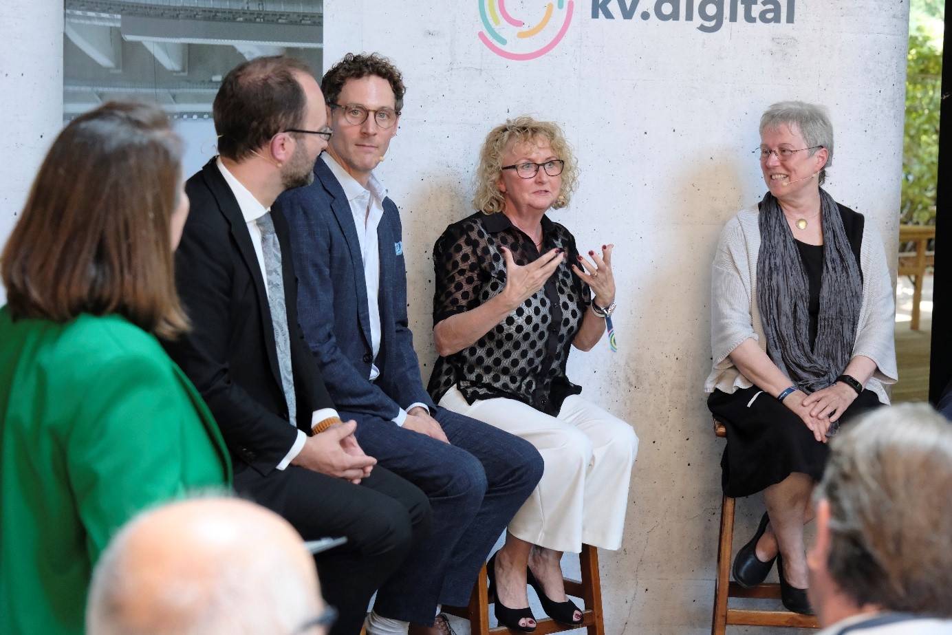 Ein Podiumsgespräch mit fünf Personen, davon drei Frauen und zwei Männer. Eine der Frauen in der Mitte gestikuliert, während sie spricht. Die anderen Personen hören aufmerksam zu und lächeln. Das Publikum ist im Vordergrund zu sehen.