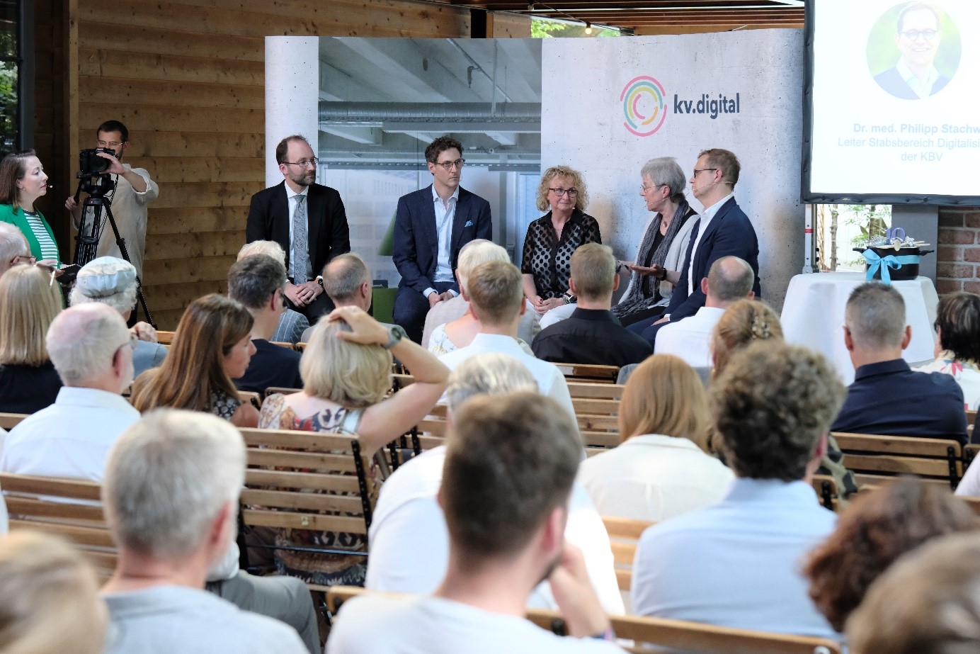 Eine Podiumsdiskussion zwischen drei Männern und zwei Frauen vor einem grauen Display mit dem Logo der kv.digital. Im Vordergrund ist das Publikum zu sehen, welches interessiert zuhört.