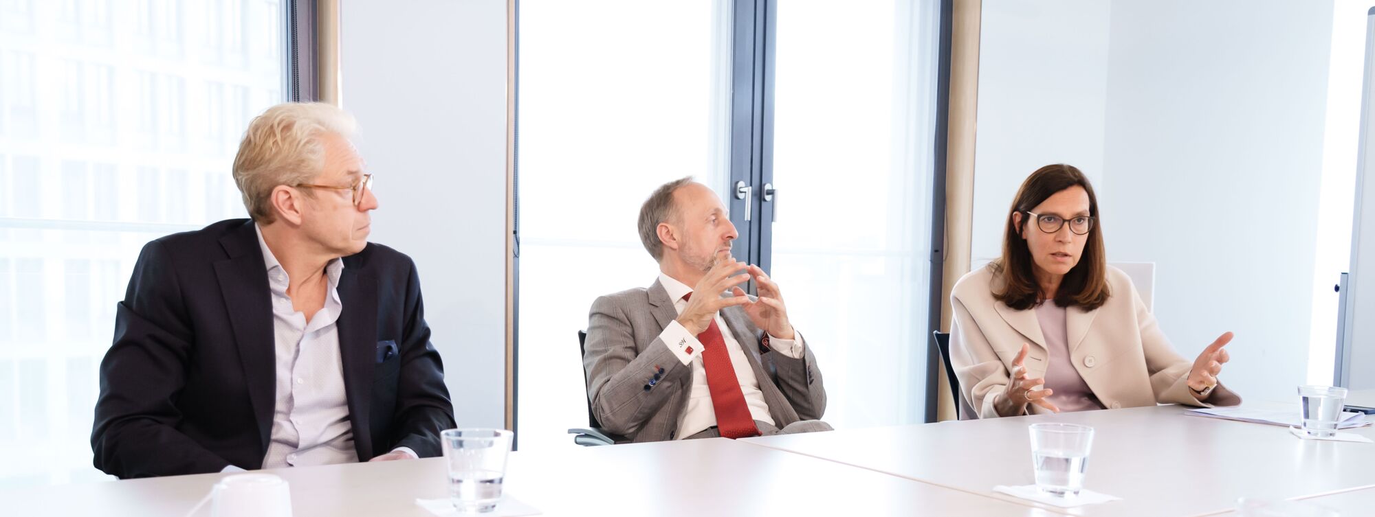 Dr. Andreas Gassen, Dr. Stephan Hofmeister und Dr. Sibylle Steiner sitzen nebeneinander an Tischen mit Wassergläser vor ihnen. Frau Steiner spricht und gestikuliert, während die beiden Herren ihr interessiert zuhören.