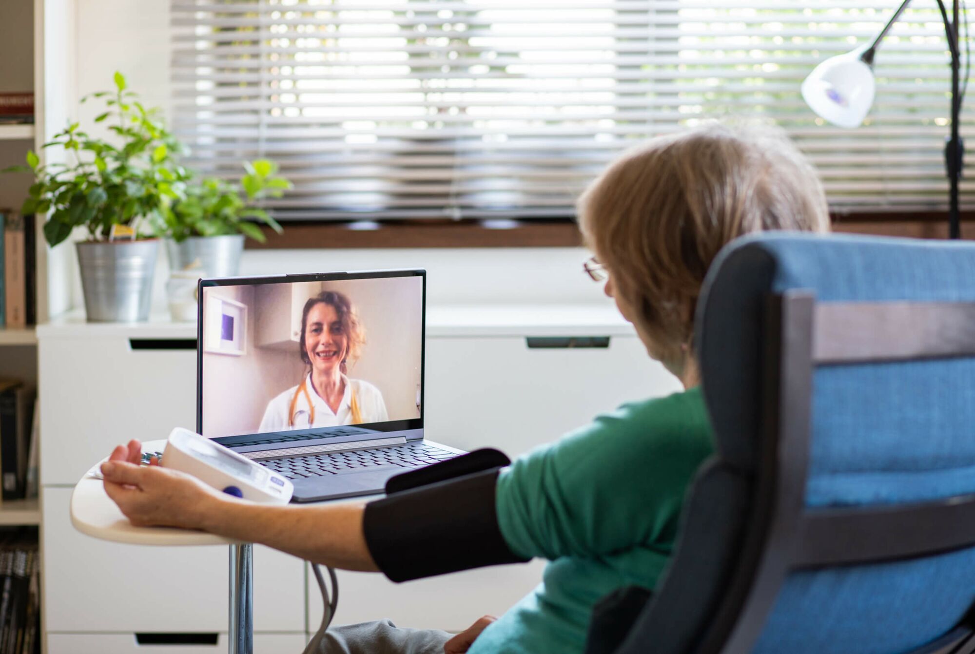 Eine ältere Dame sitzt in einem Sessel vor einem kleinen Tisch mit einem Laptop drauf. Sie hat die Manschette des Pulsmessgerätes an ihrem linken Arm und ist im Gespräch mit einer Ärztin via Videocall.